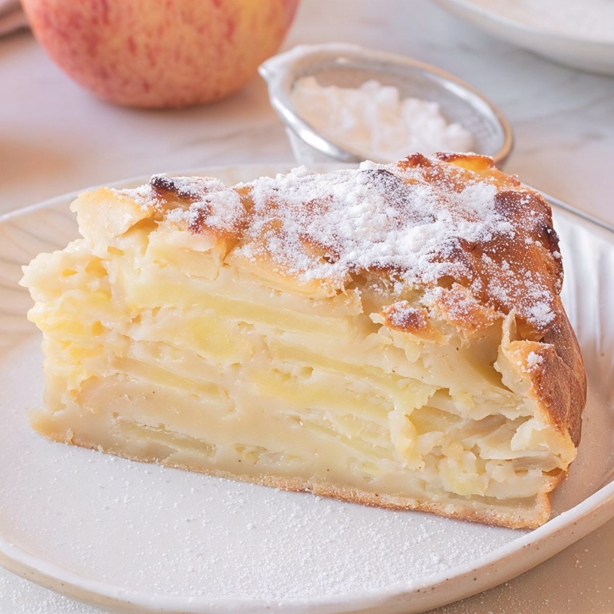 A slice of French invisible apple cake displaying thin, tender layers of sliced apples held together by a light custard batter and dusted with powdered sugar on top