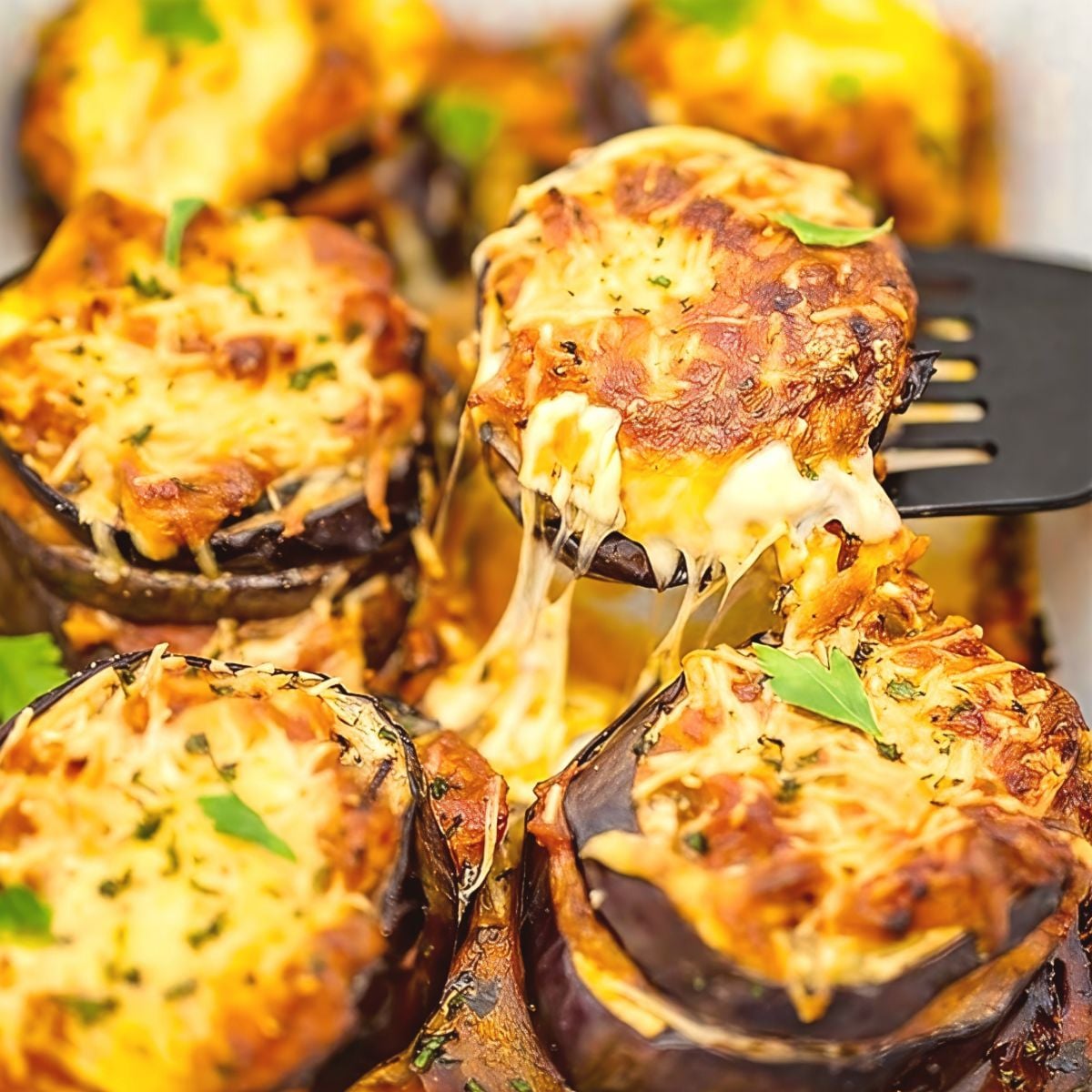 A spatula lifts a piece of cheesy keto eggplant parmesan with melted mozzarella pulls and fresh parsley garnish from a baking dish, making a delicious addition to keto thanksgiving sides.