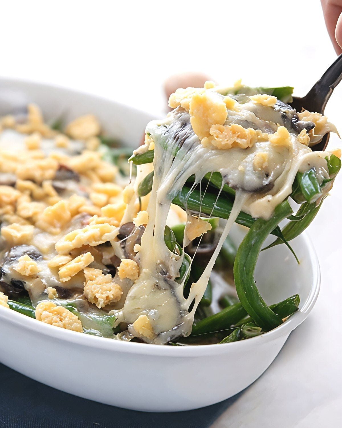 A serving utensil lifts a scoop of creamy green bean casserole with melted cheese pulls, mushrooms, and crispy toppings from a white baking dish, perfect for keto thanksgiving sides.