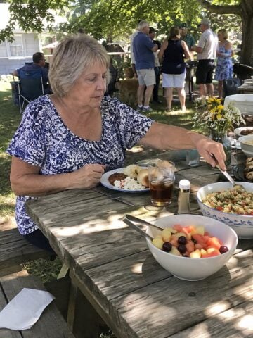 At a family reunion picnic table in mid-afternoon, a woman in her sixties sits on her own, reaching for a serving spoon beside a half-filled plate while relatives gather in the background.