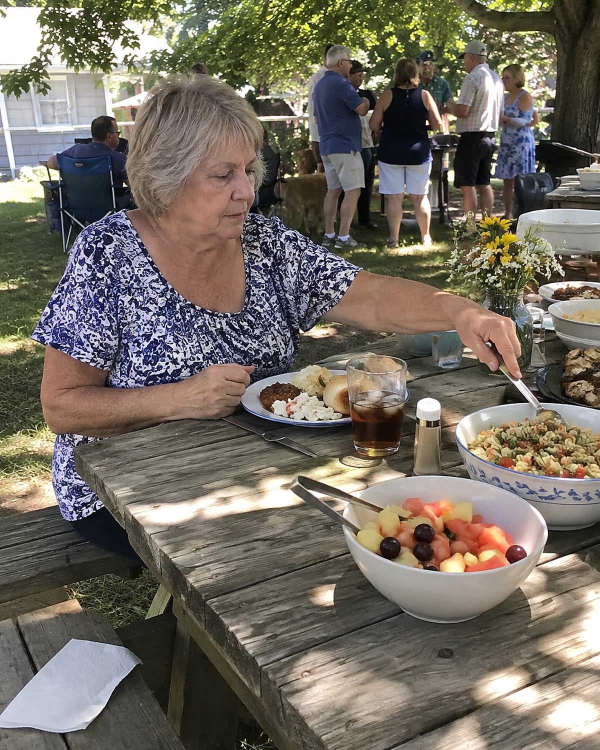 At a family reunion picnic table in mid-afternoon, a woman in her sixties sits on her own, reaching for a serving spoon beside a half-filled plate while relatives gather in the background.