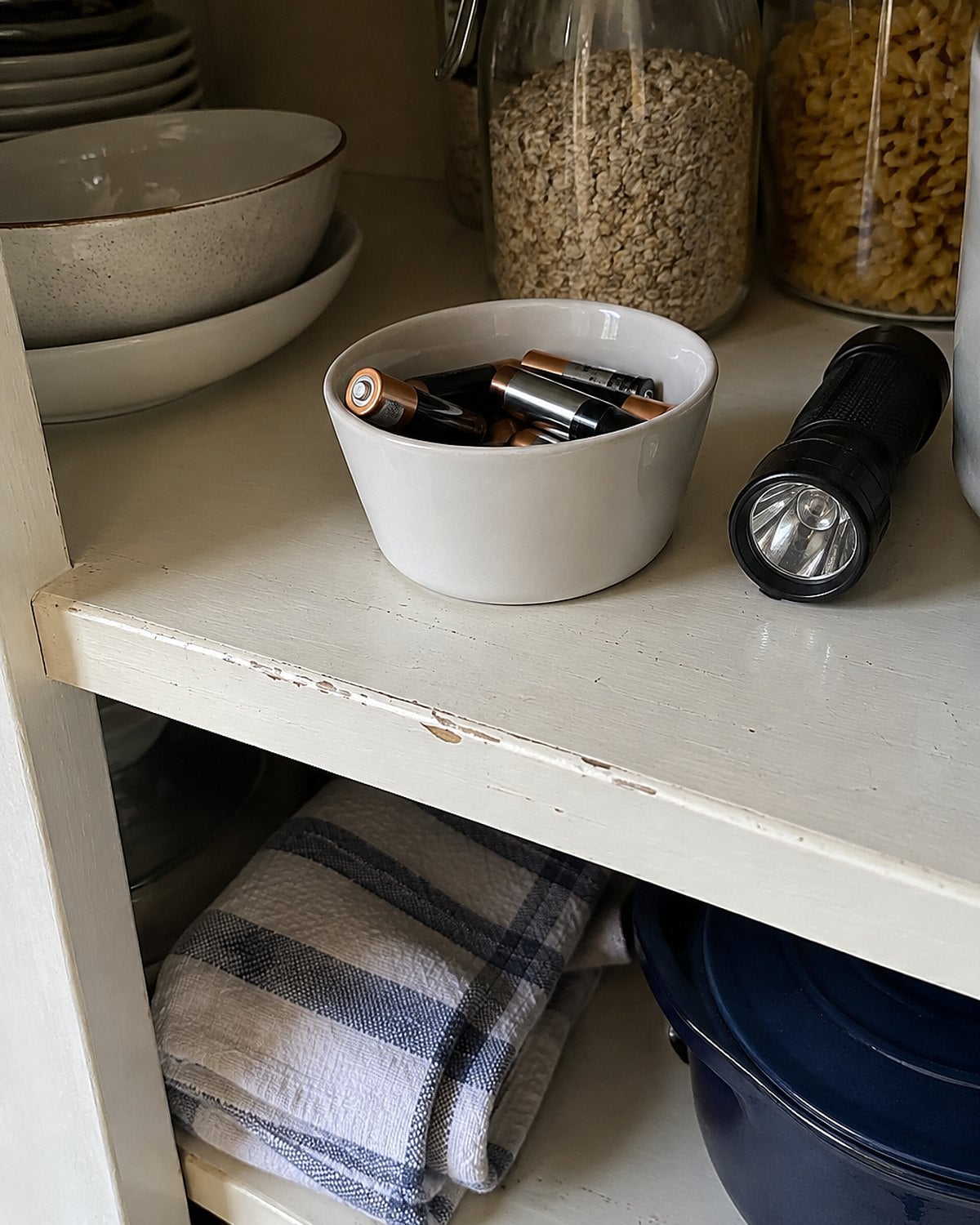 Used batteries in a small container beside a flashlight on a pantry shelf.