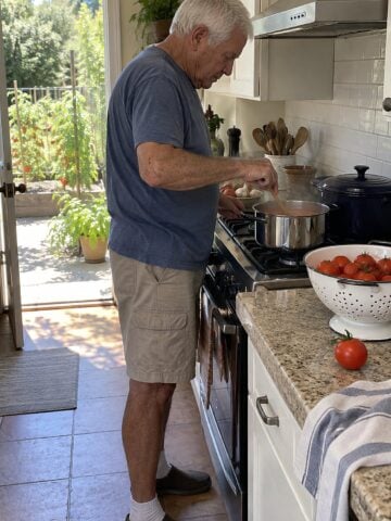 At a garden bed of tomatoes around noon, a man in his seventies stands at the stove stirring a pot beside a colander of picked tomatoes, mid-moment and unposed.