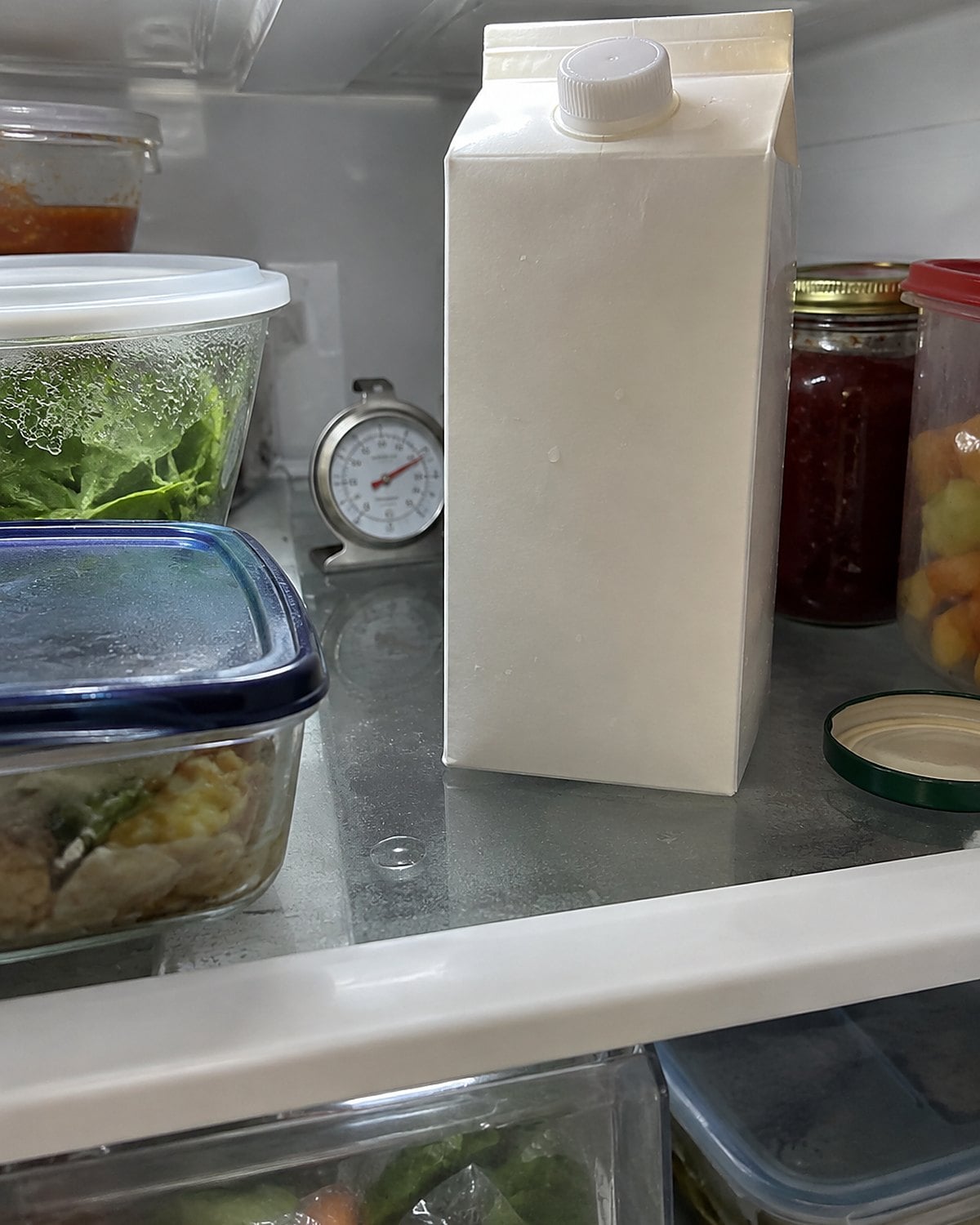 A small thermometer sits behind a milk carton on the middle shelf of a refrigerator.