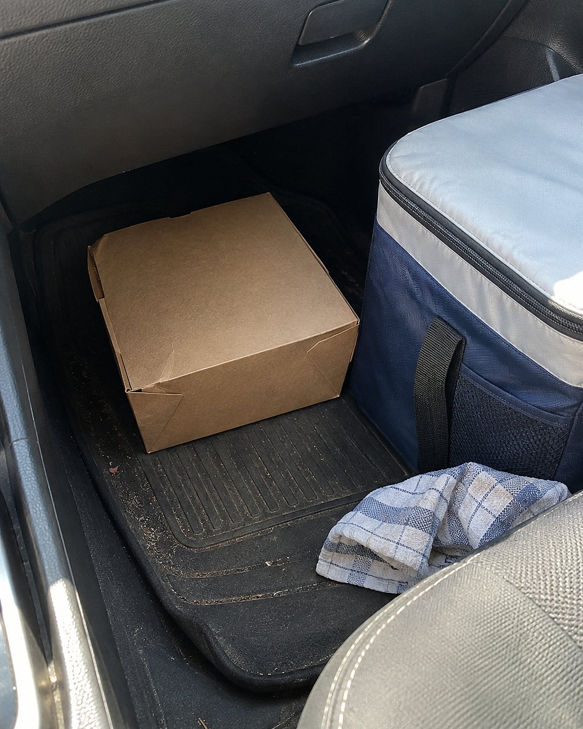 A closed takeout carton sits beside an insulated cooler in the passenger footwell of a car.