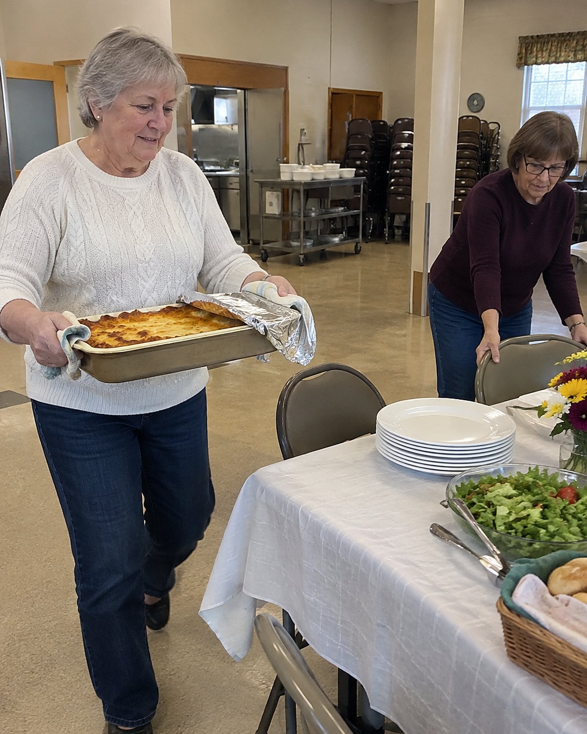 Hands carry a pan of baked pasta past stacked chairs in a church hall.
