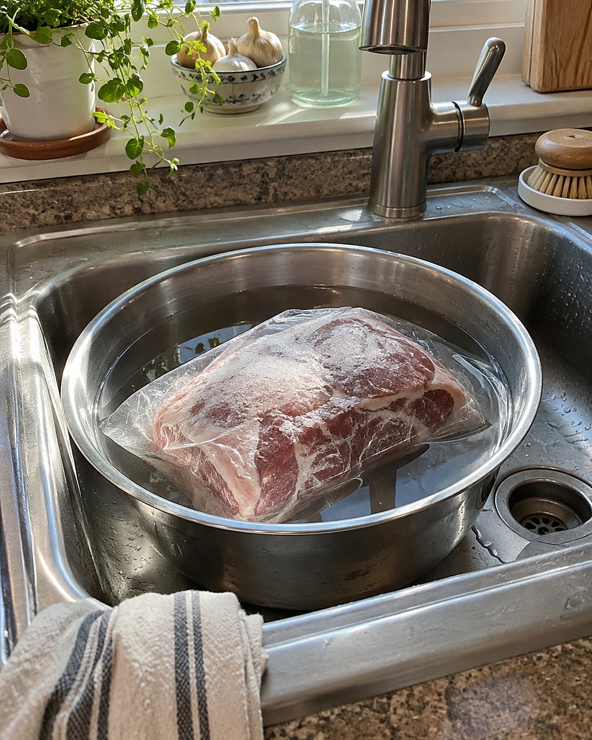 A wrapped frozen roast sits in a bowl of cold water in the kitchen sink.