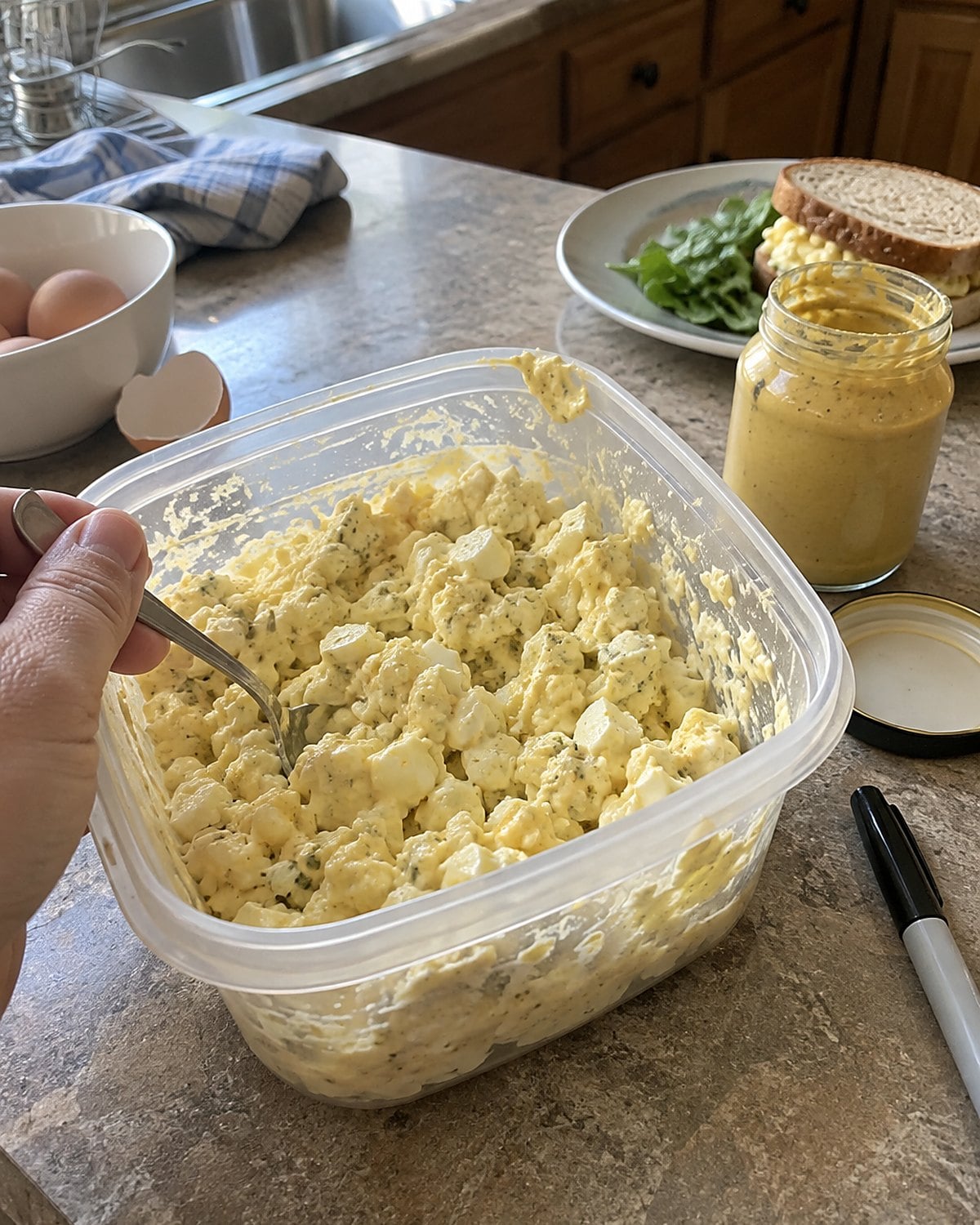 An opened container of egg salad sits on the counter beside mustard and a marker.