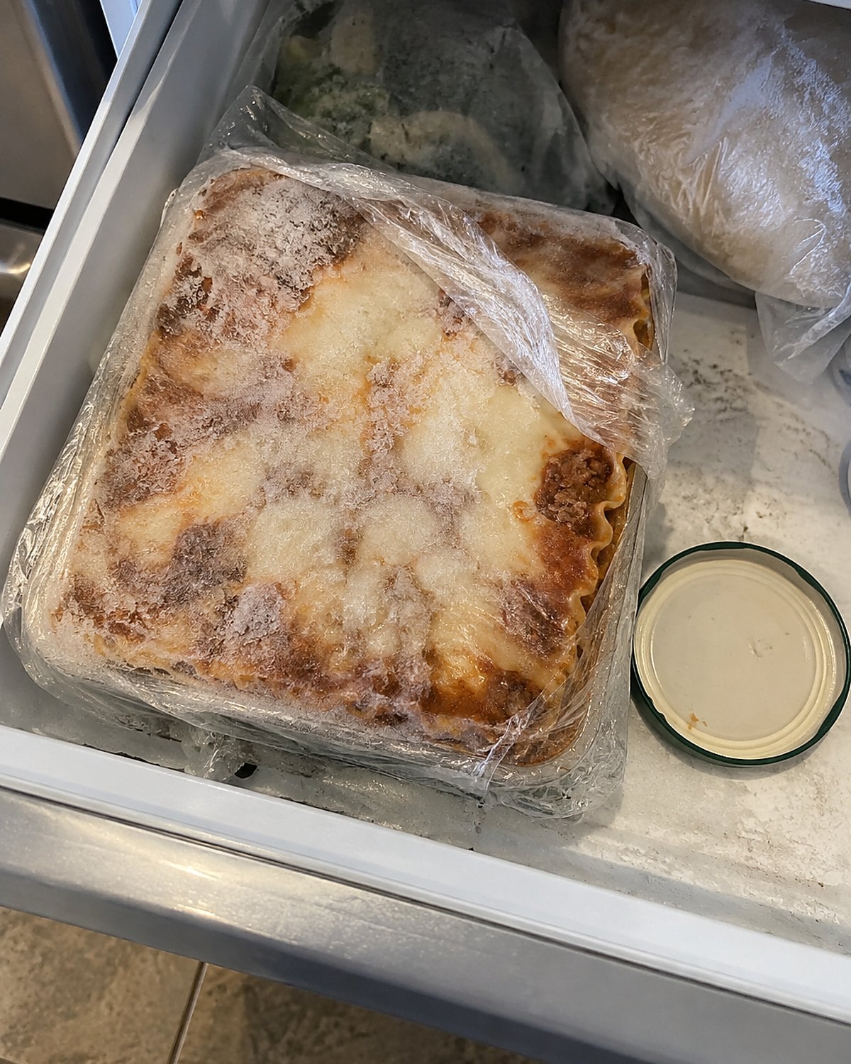 A frost-marked square of wrapped lasagna sits in an open freezer drawer.
