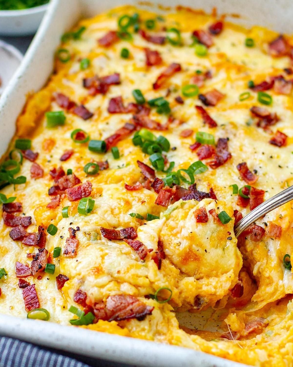 A metal spoon scoops into a casserole dish of creamy loaded mashed potatoes topped with melted cheddar cheese, crispy bacon bits, and sliced green onions.