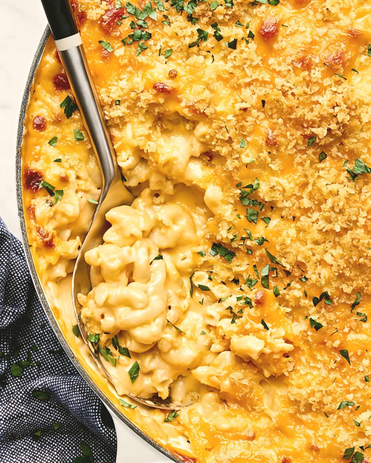 A metal spoon scoops up a creamy portion of classic homemade macaroni and cheese topped with a crispy, golden-brown breadcrumb topping and chopped herbs