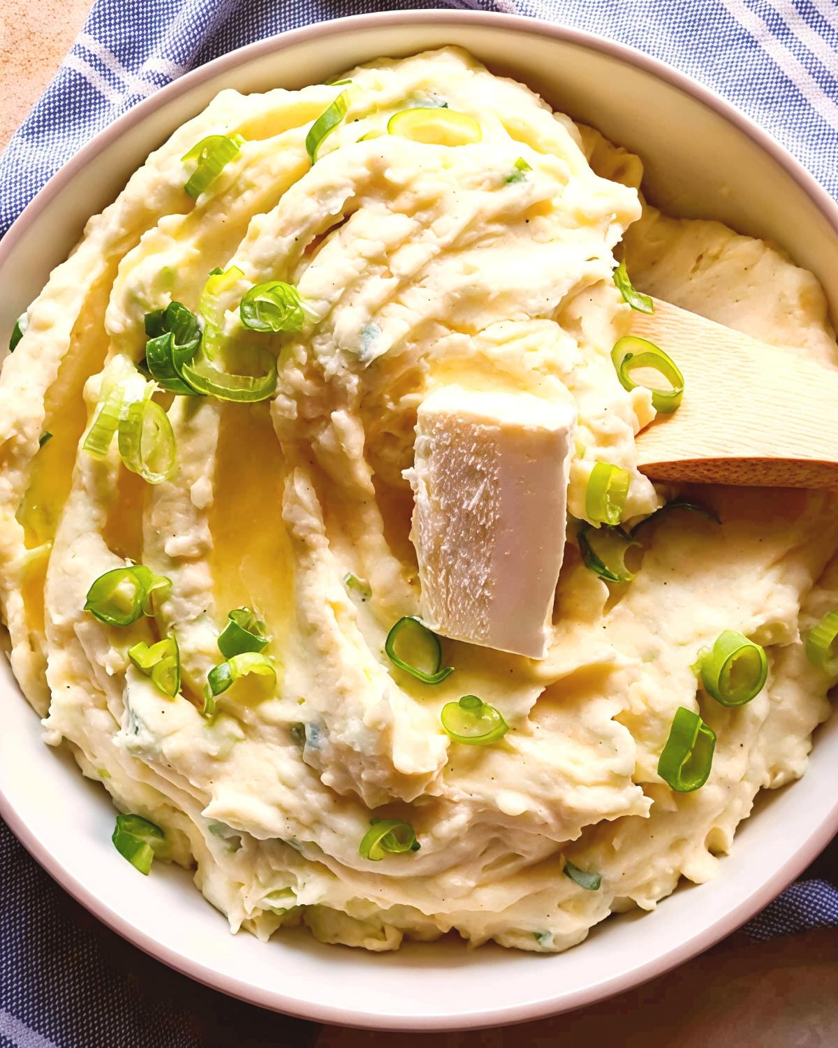 A bowl of creamy, swirled mashed potatoes is topped with melted butter, green onions, and a large slice of butter, being served with a wooden spoon.