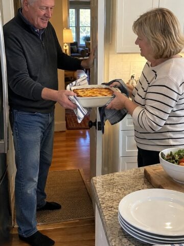 A couple in their sixties at a neighbor's kitchen doorway just before dinner, one handing over a warm casserole dish while the other holds the door.
