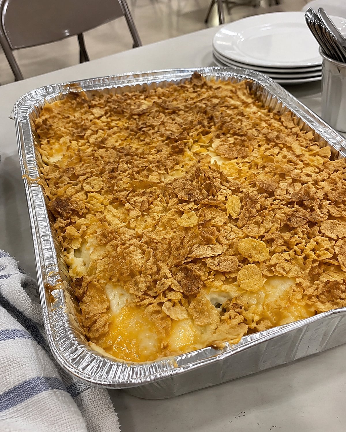 A foil baking dish of funeral potatoes with uneven crushed cornflakes on a church hall table.