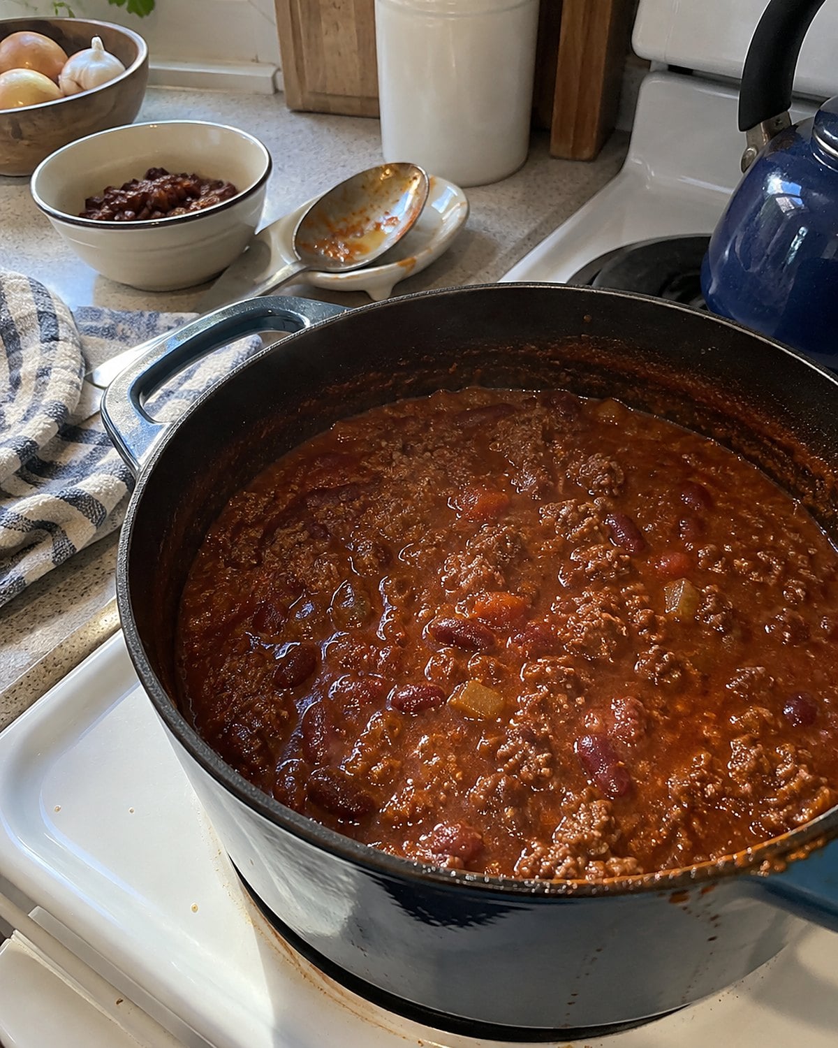 A dark pot of chili simmering on a white stove, with a spoon resting beside a small bowl of beans.