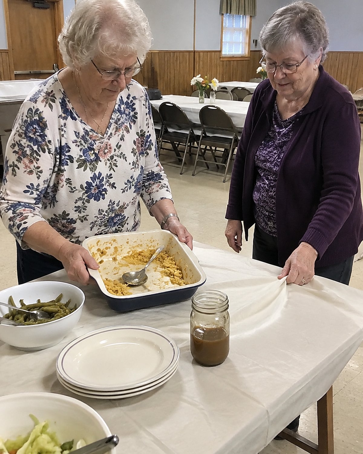 Hands collect a partly emptied casserole dish from a church hall serving table.