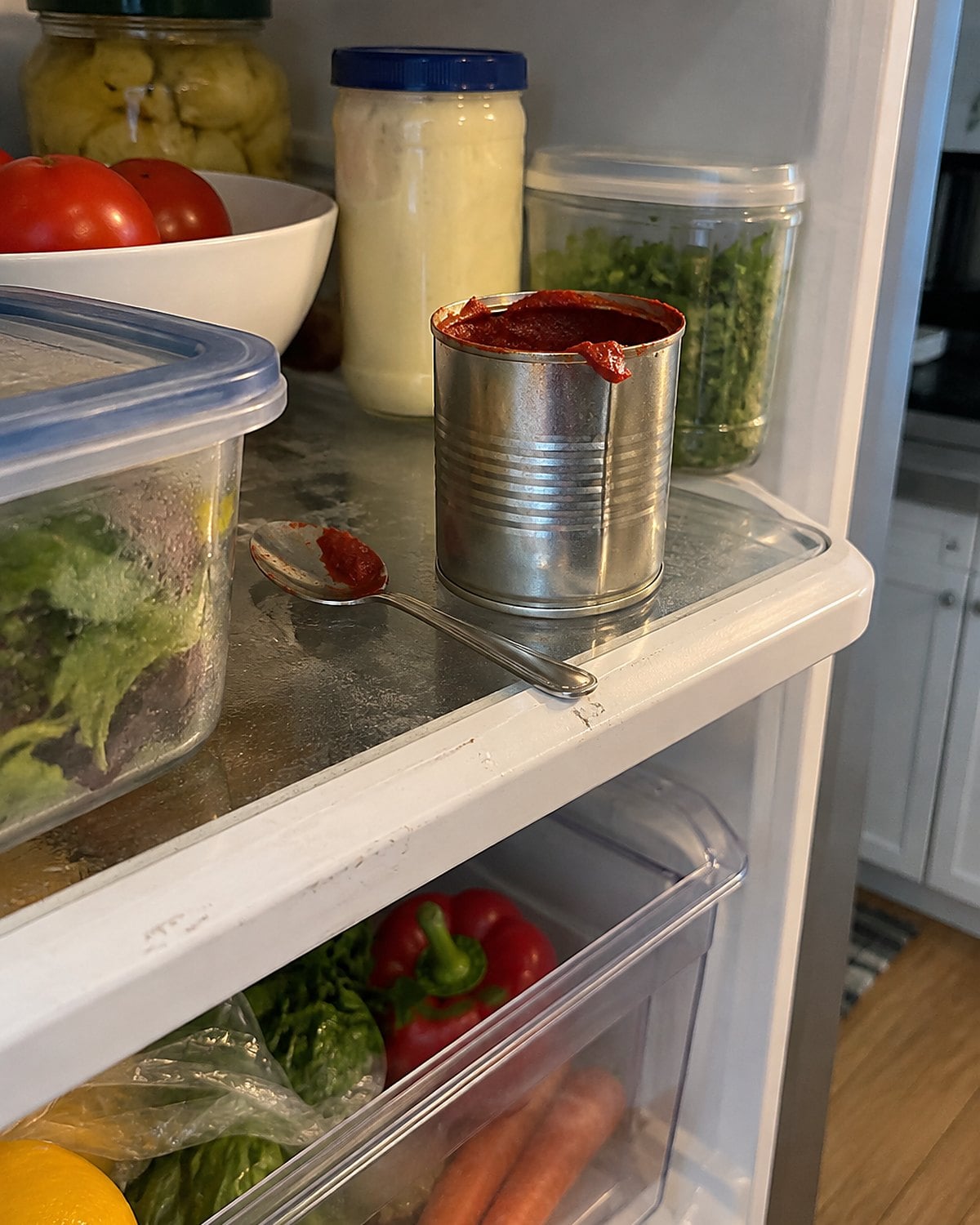 An opened can of tomato paste and a spoon sit on a crowded refrigerator shelf.