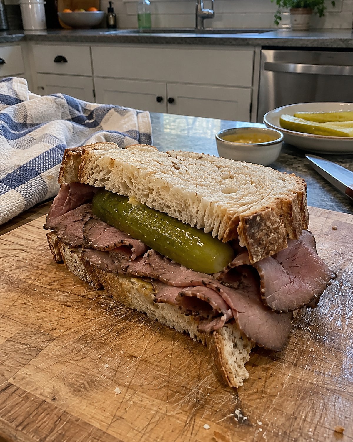 Thin roast beef and a pickle rest between uneven slices of bread on a kitchen cutting board.