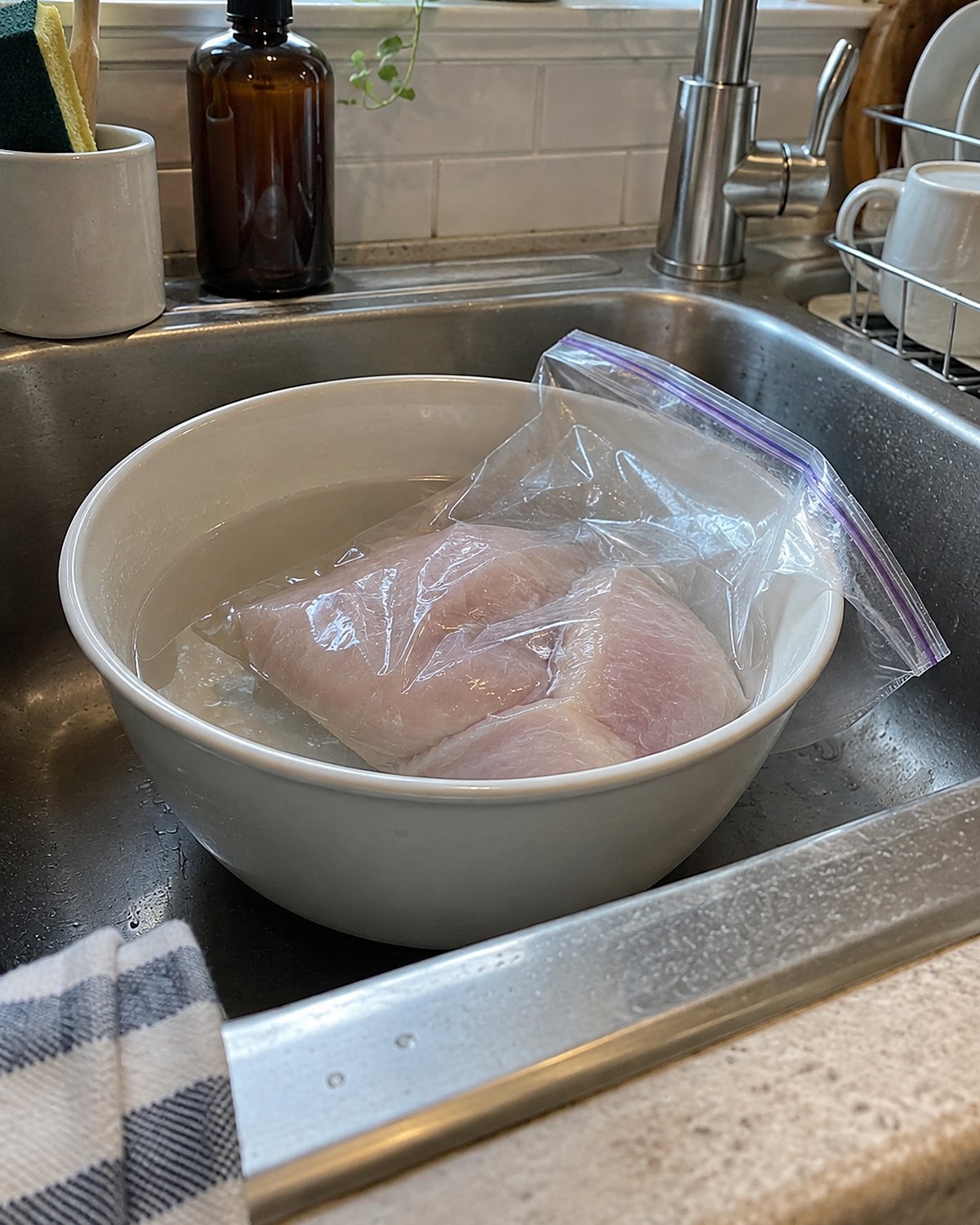 A sealed bag of thawing chicken sits in a bowl of cold water in a clean sink.