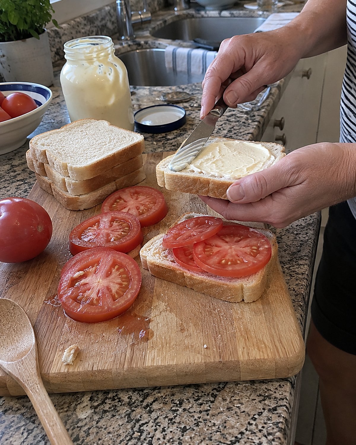 Tomato slices and soft white bread wait on a cutting board beside a jar of mayonnaise.