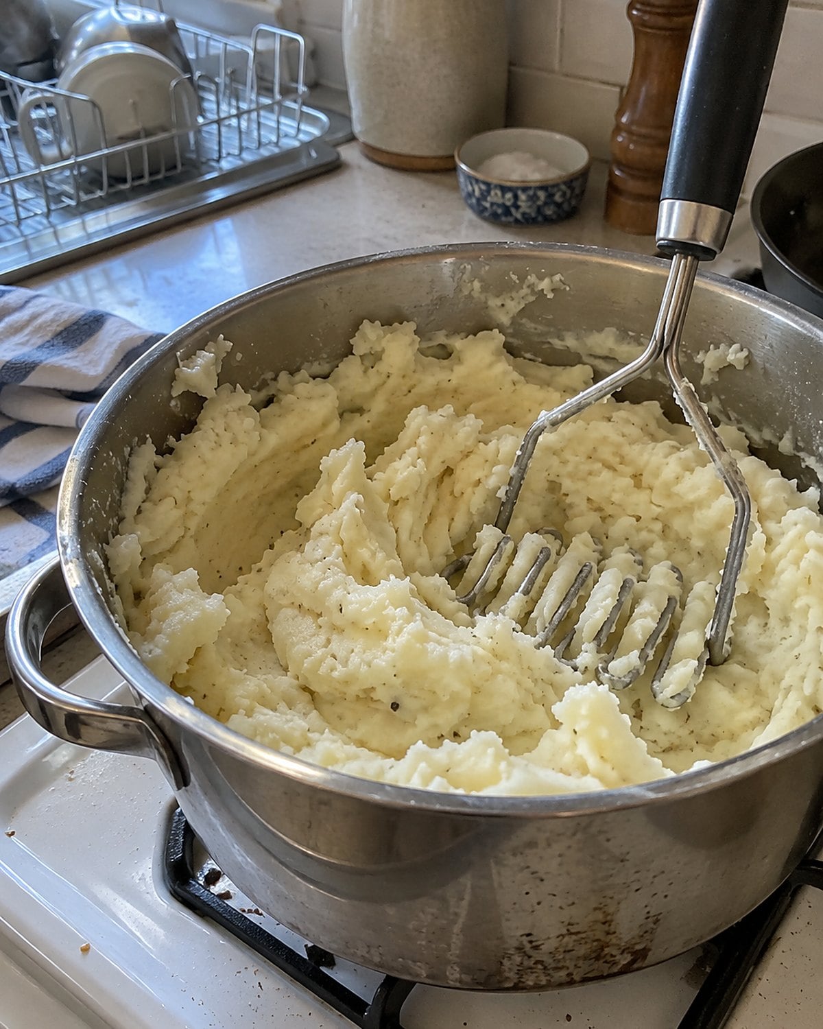 A potato masher stands in a pot of mashed potatoes with soft ridges across the surface.