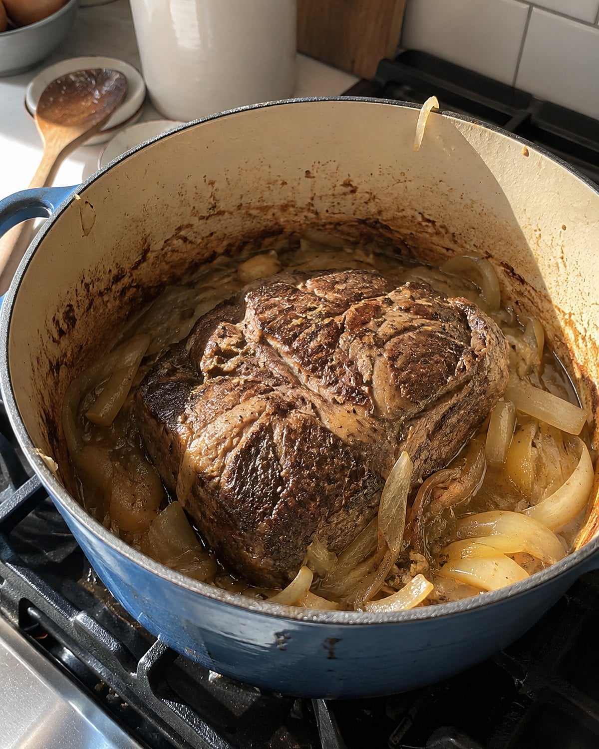 A chuck roast rests among slumped onions in an open Dutch oven on the stove.
