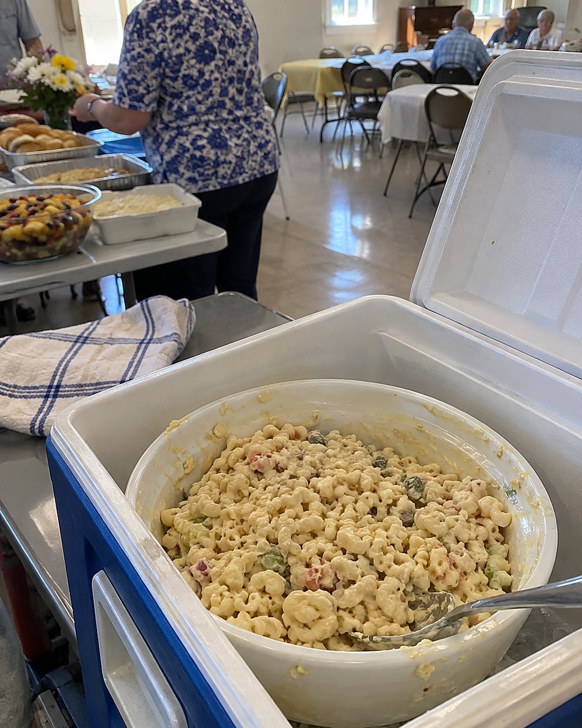A bowl of macaroni salad sits in a cooler beside a church hall serving table.