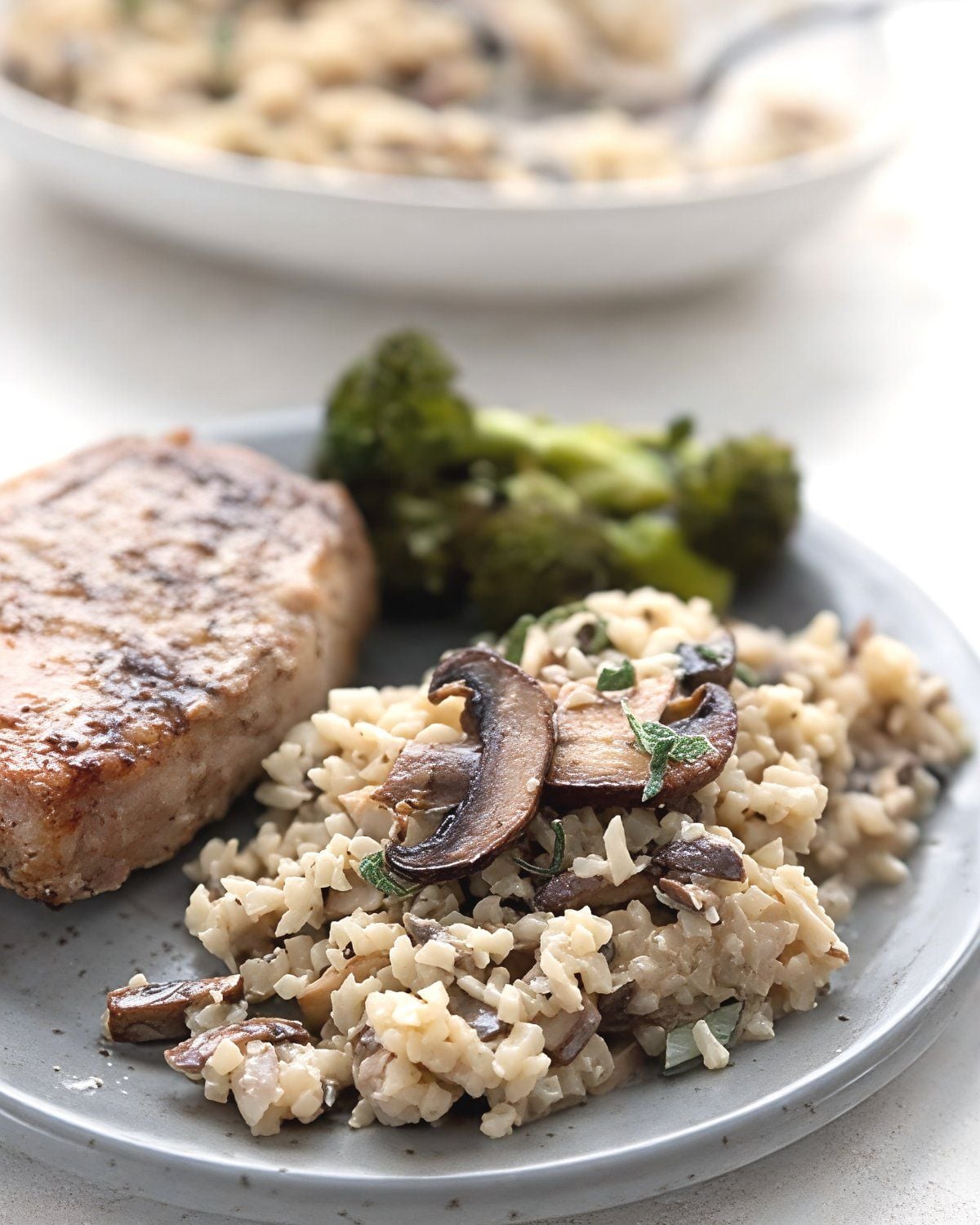 Creamy mushroom cauliflower risotto garnished with sliced sautรฉed mushrooms and fresh sage leaves is served alongside a roasted pork chop and broccoli for flavorful keto thanksgiving sides.