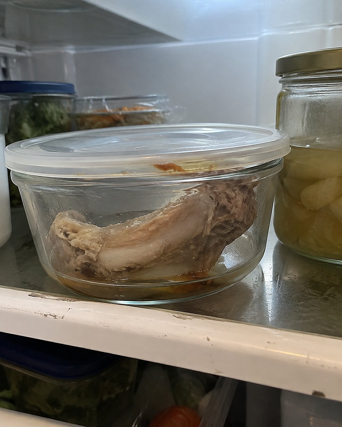 A chicken neck rests in a covered glass container on a refrigerator shelf.