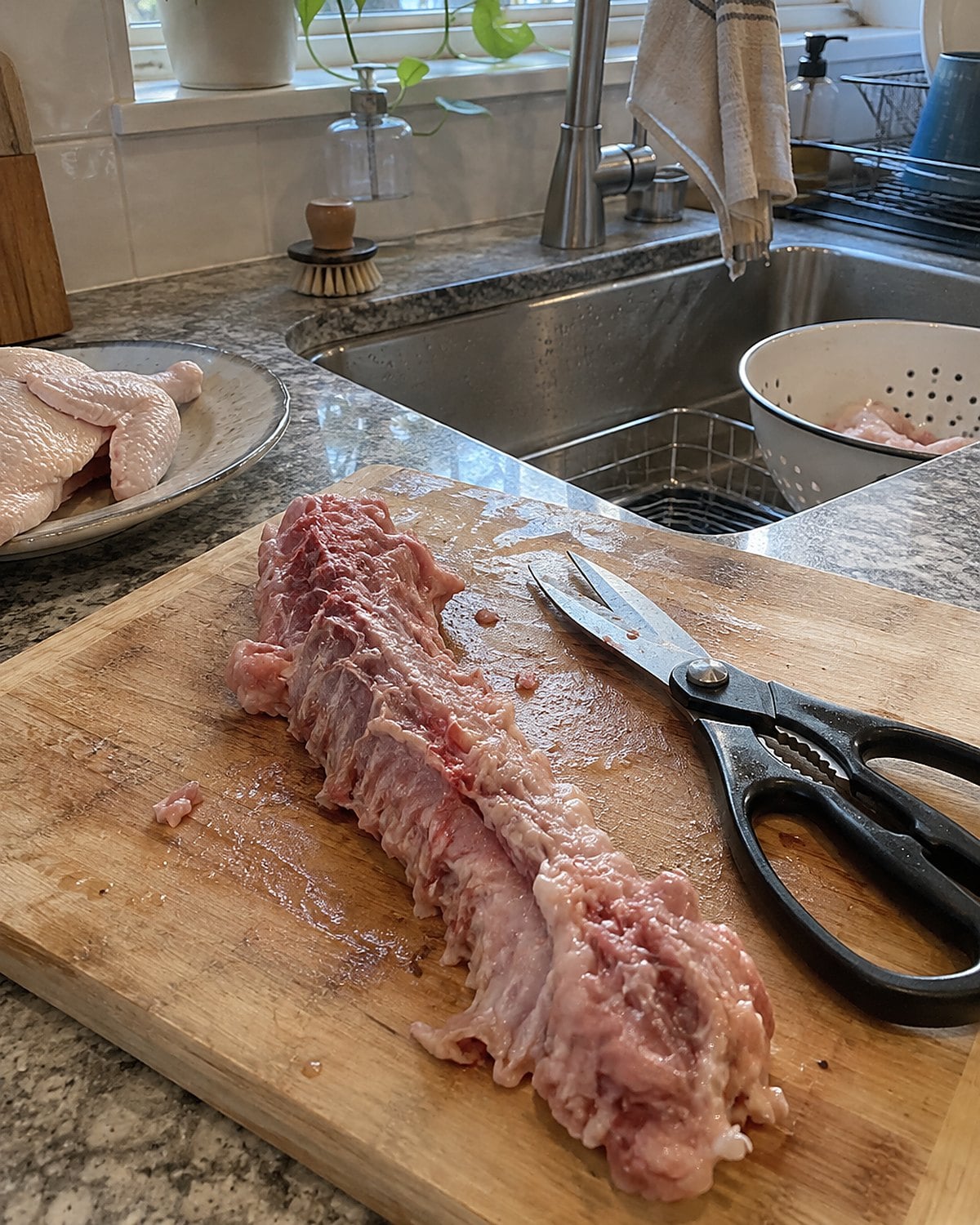 Kitchen shears lie beside a raw chicken backbone on a worn cutting board near the sink.