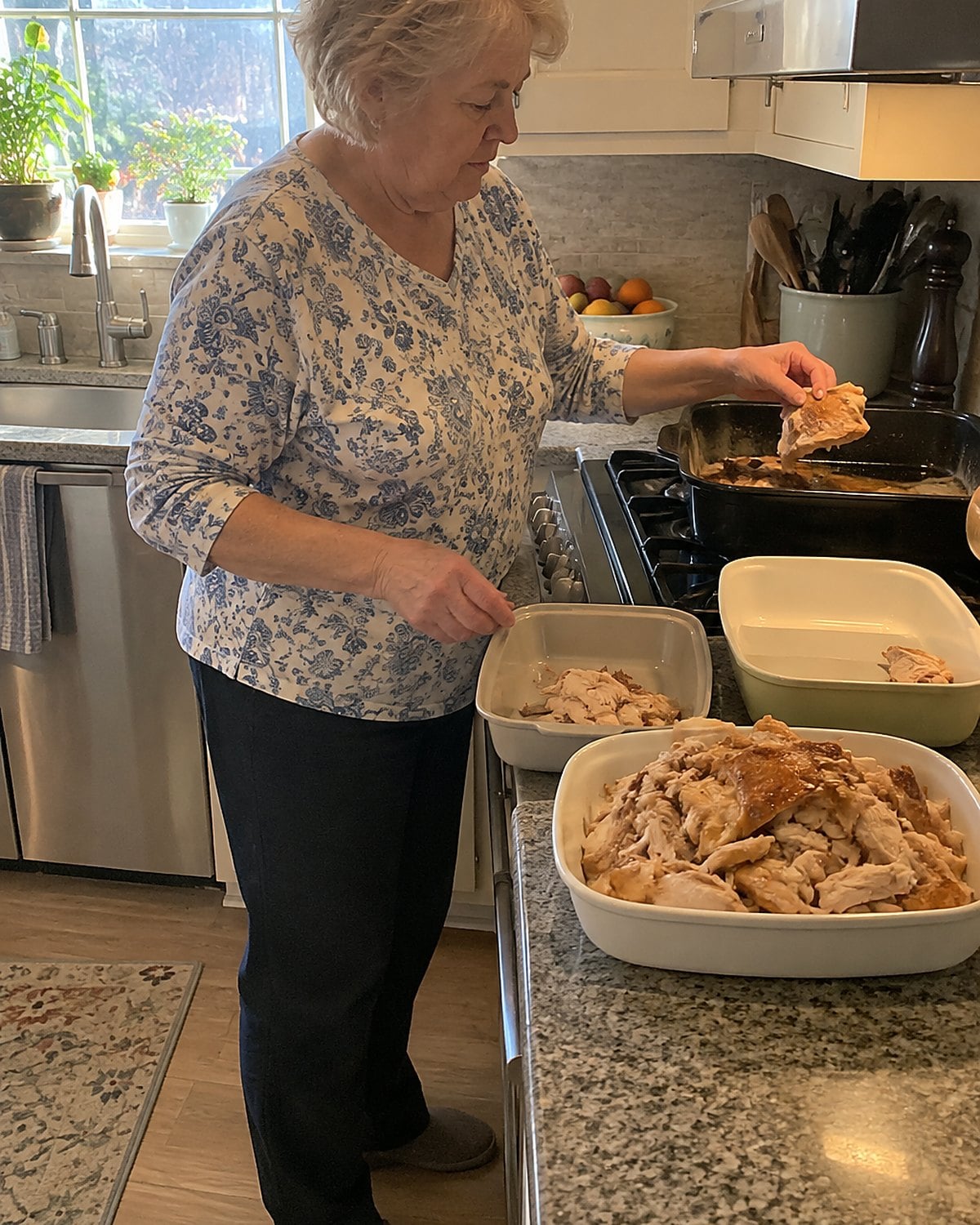 An older woman's hands divide roast chicken among shallow containers beside the stove.