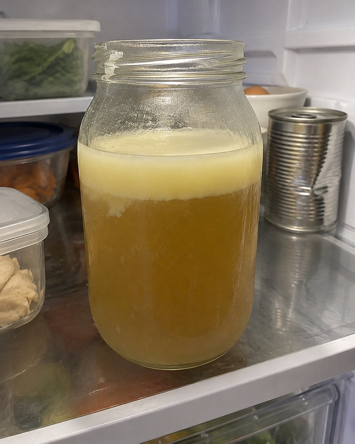 A jar of chilled broth with a layer of fat sits on a refrigerator shelf.