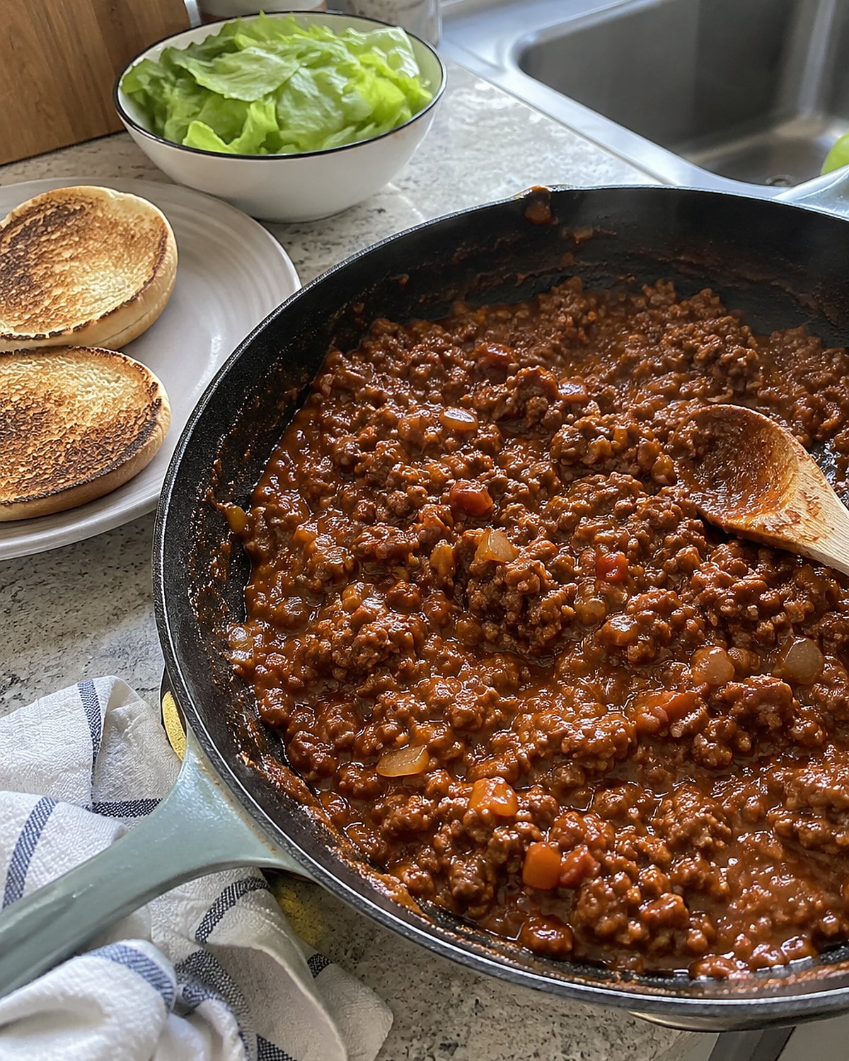A toasted bun on a counter beside a skillet of thick sloppy Joe filling.