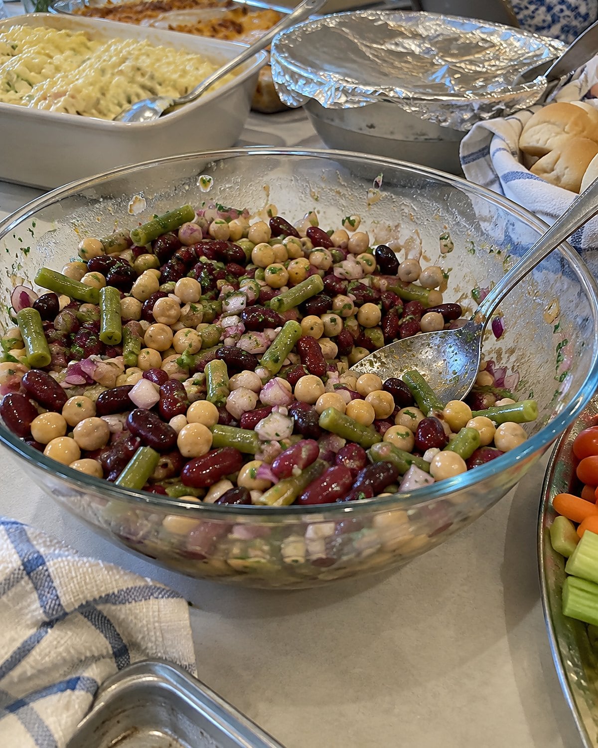 A bowl of three-bean salad among covered dishes on a church hall table.