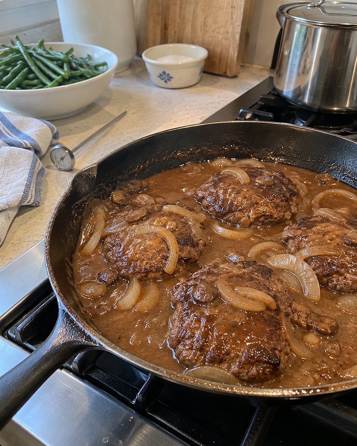 Salisbury steak patties in a skillet of onion gravy, with a thermometer beside the stove.