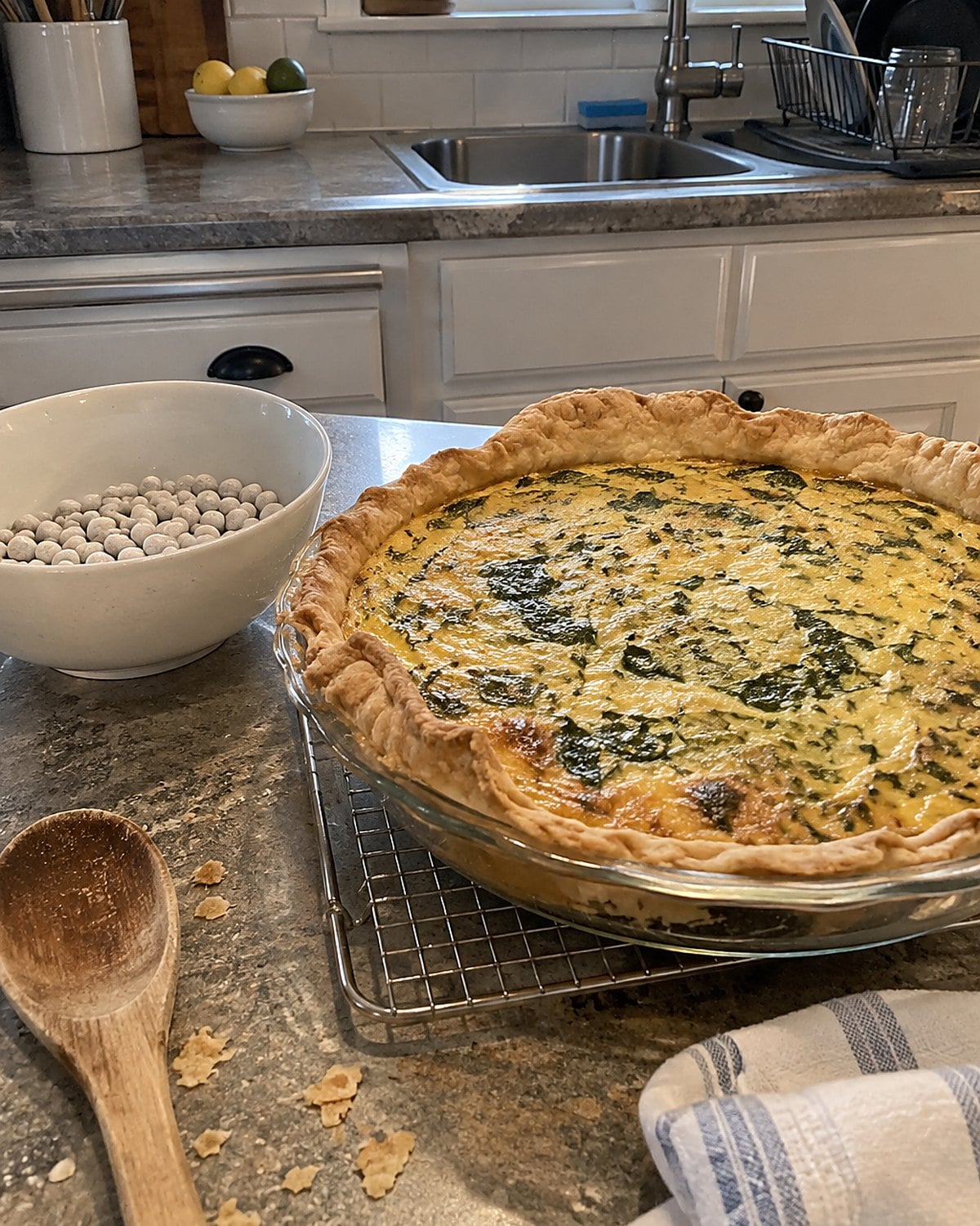 A quiche cooling beside its empty pie weights on a kitchen counter, one edge of crust slightly uneven.