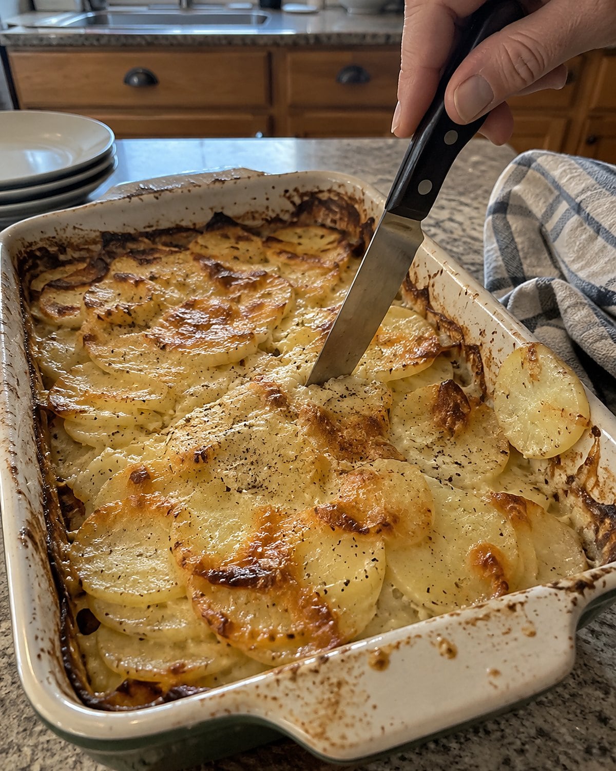 A knife tip pressed into the center of scalloped potatoes in a worn baking dish.