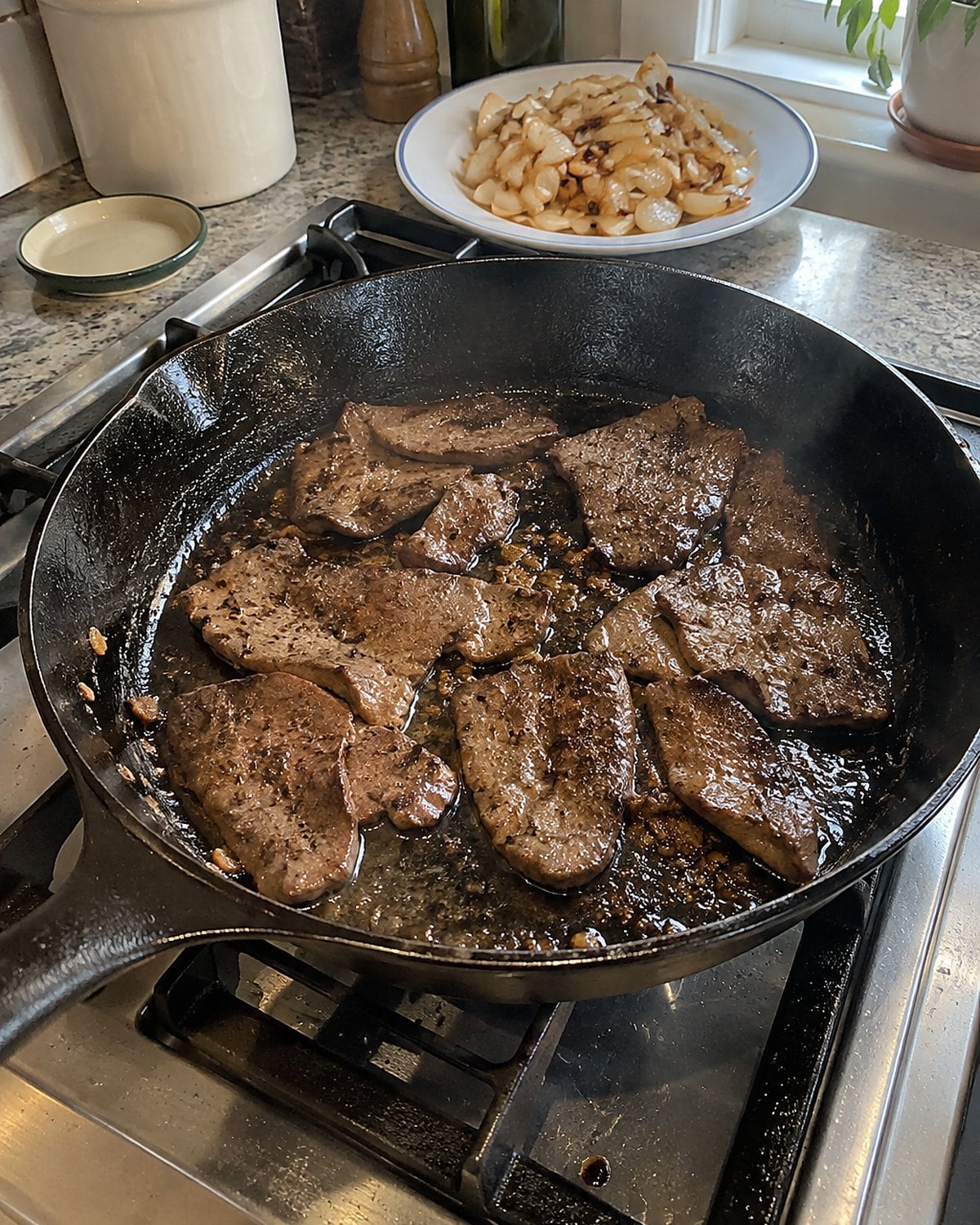 Thin pieces of liver browning in a cast-iron skillet, with cooked onions waiting on a nearby plate.