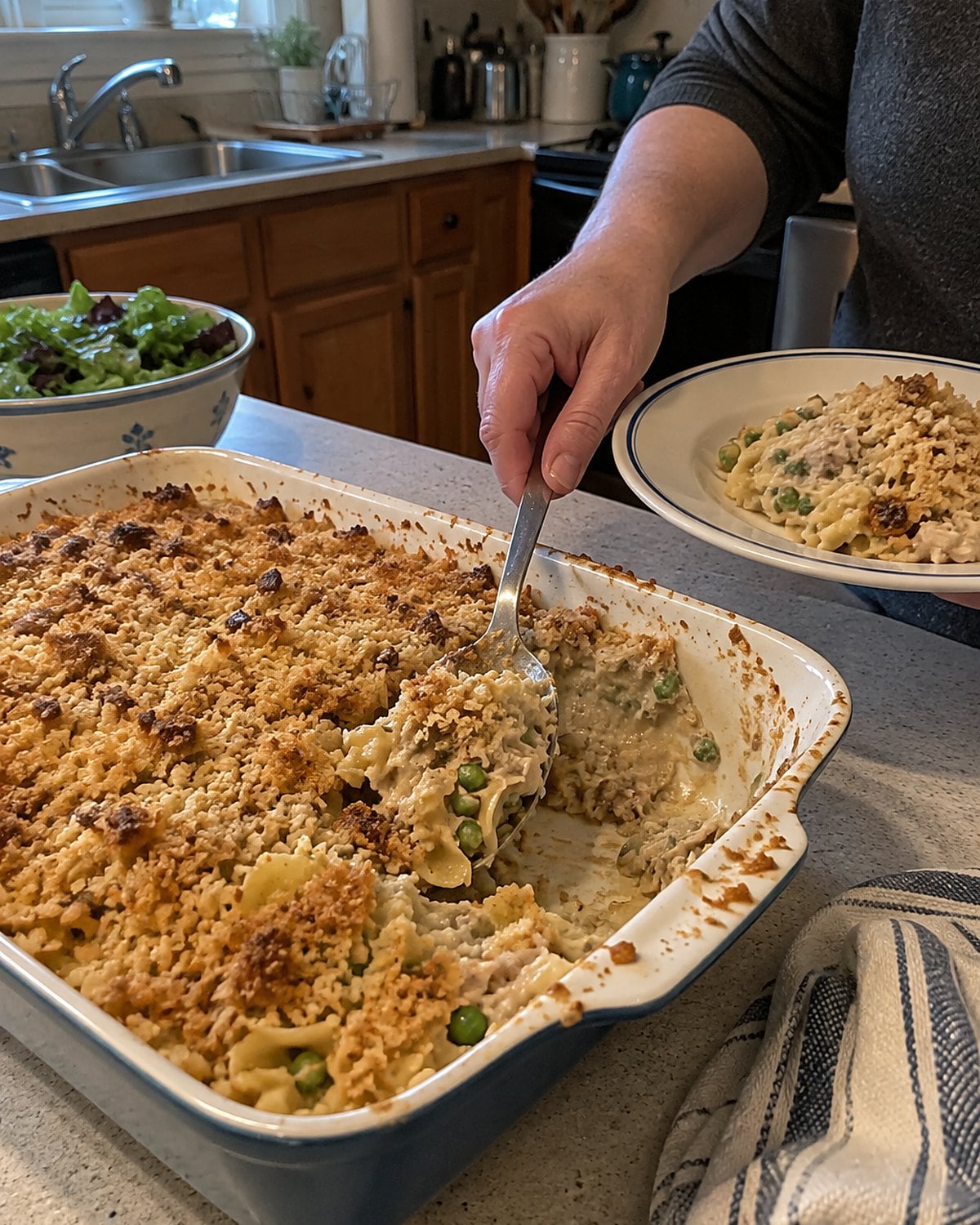 A spoon breaking the crumb topping of tuna noodle casserole in a chipped baking dish.