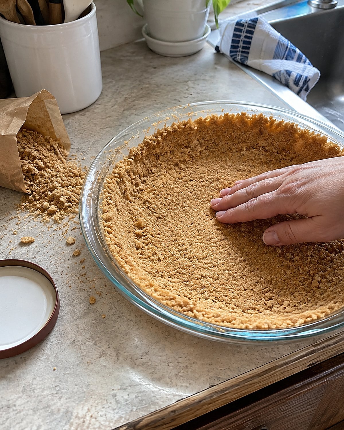 Crushed graham crackers spill from a sleeve beside a buttered pie plate in the sink-side work area.
