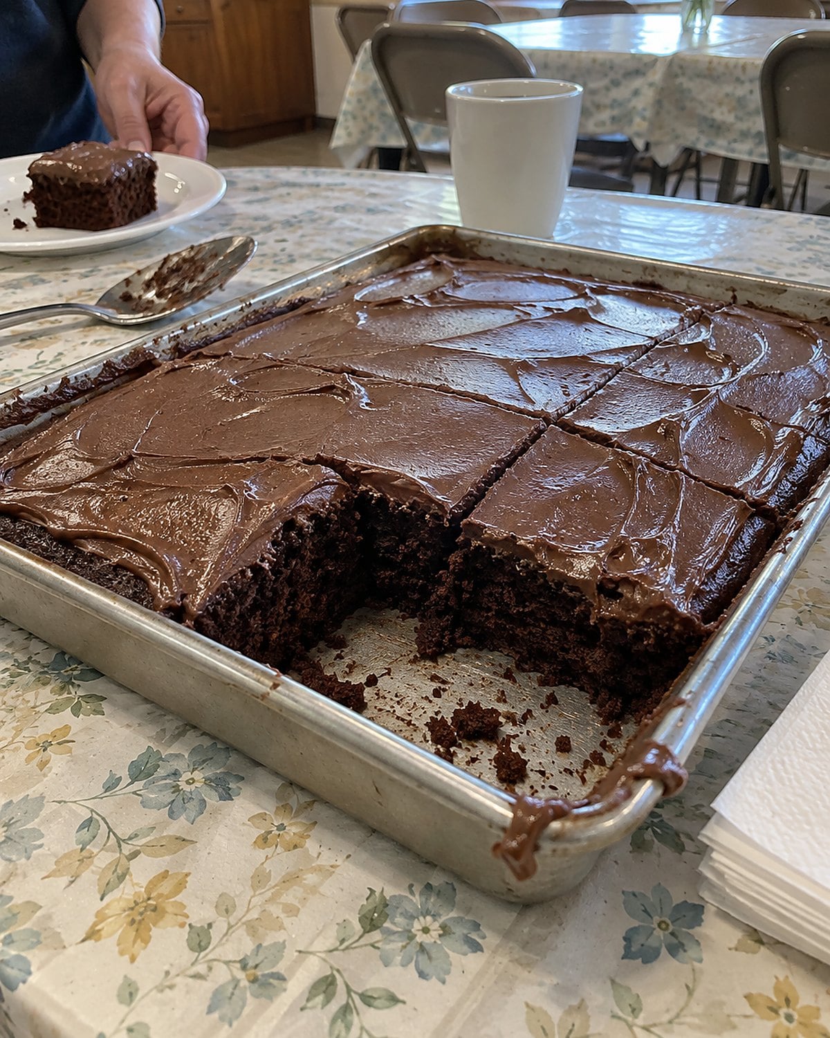 A pan of chocolate sheet cake with an unevenly cut corner sits beside paper napkins on a church hall table.