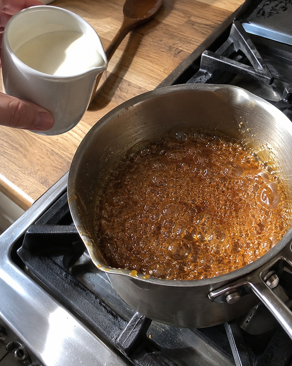 Amber caramel bubbles in a small saucepan on the stove beside a ready cup of cream.