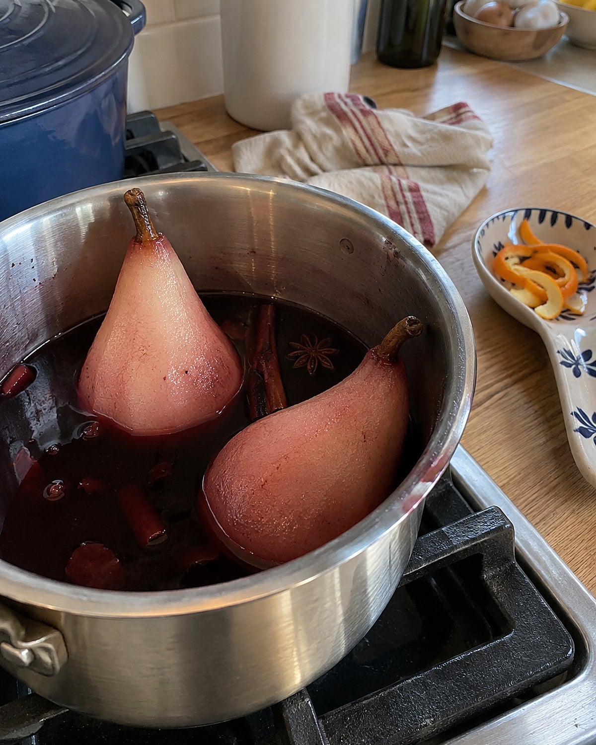 Two peeled pears stand in red wine in a saucepan, with orange peel on a spoon rest nearby.