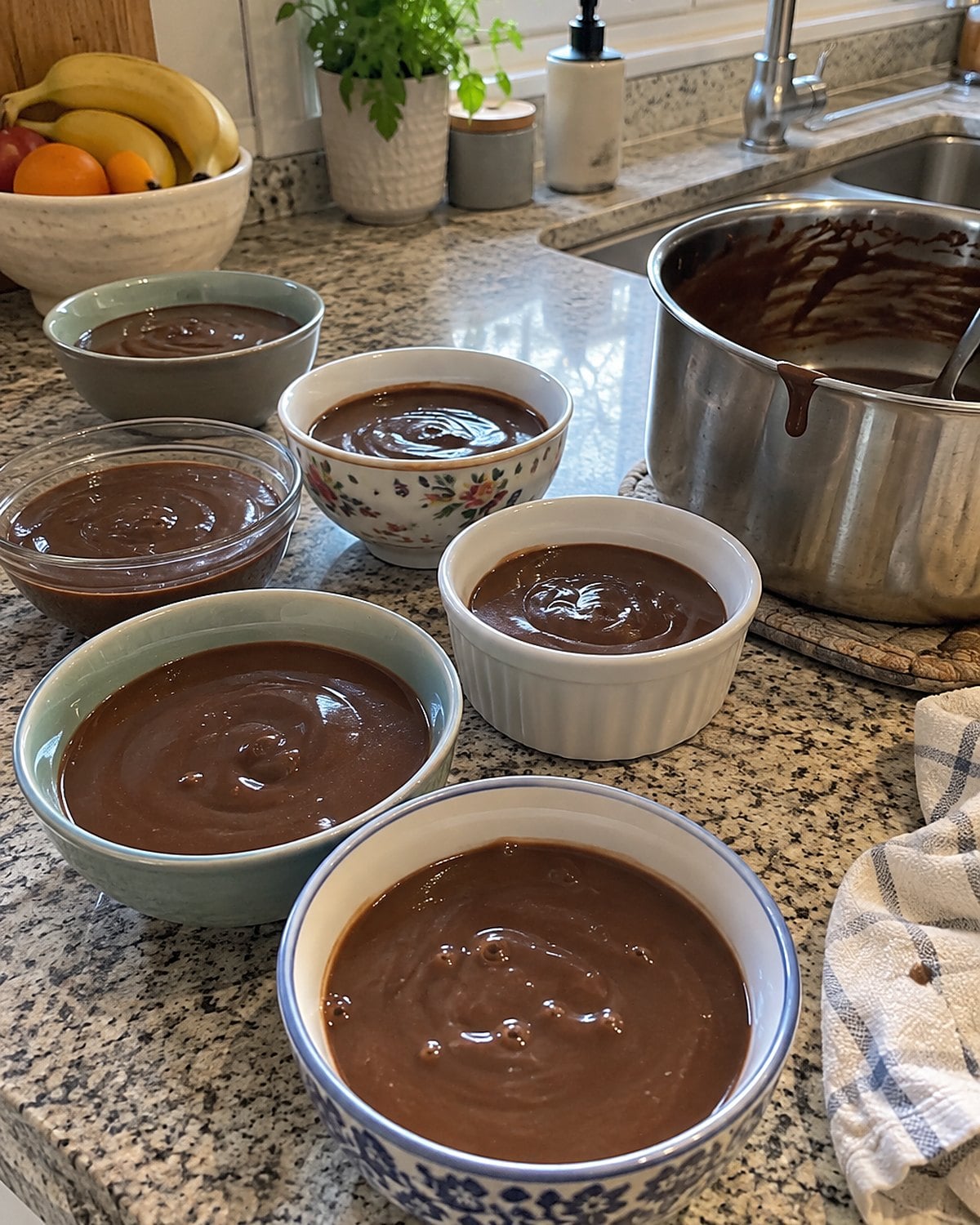 Small mismatched bowls of chocolate pudding cool on a counter beside a chocolate-streaked saucepan.
