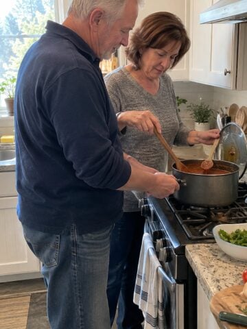 At a stove around noon, a couple in their sixties stir a pot together while its lid rests propped on a spoon, neither looking at the camera.