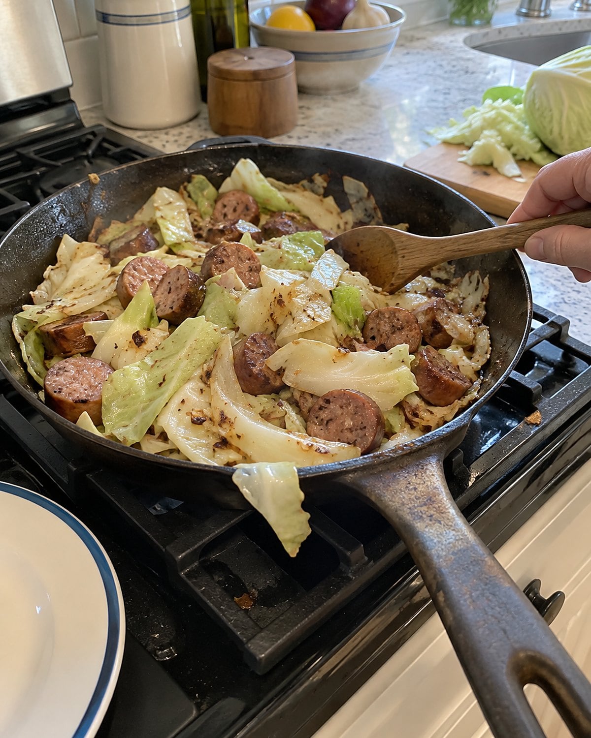 Wide cabbage strips and browned sausage fill a skillet beside a wooden spoon.