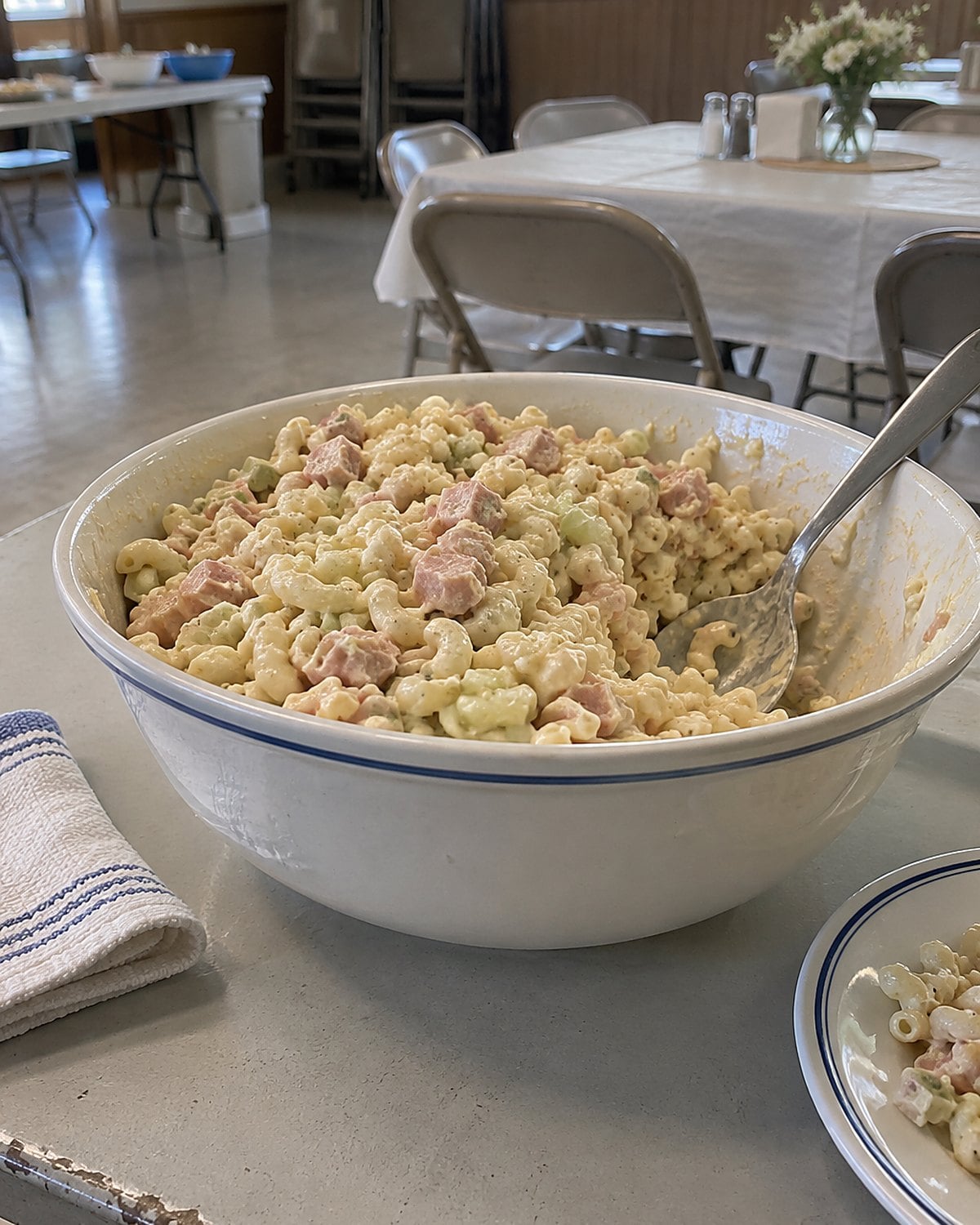 A bowl of macaroni salad with chopped bologna and celery sits on a church hall table.