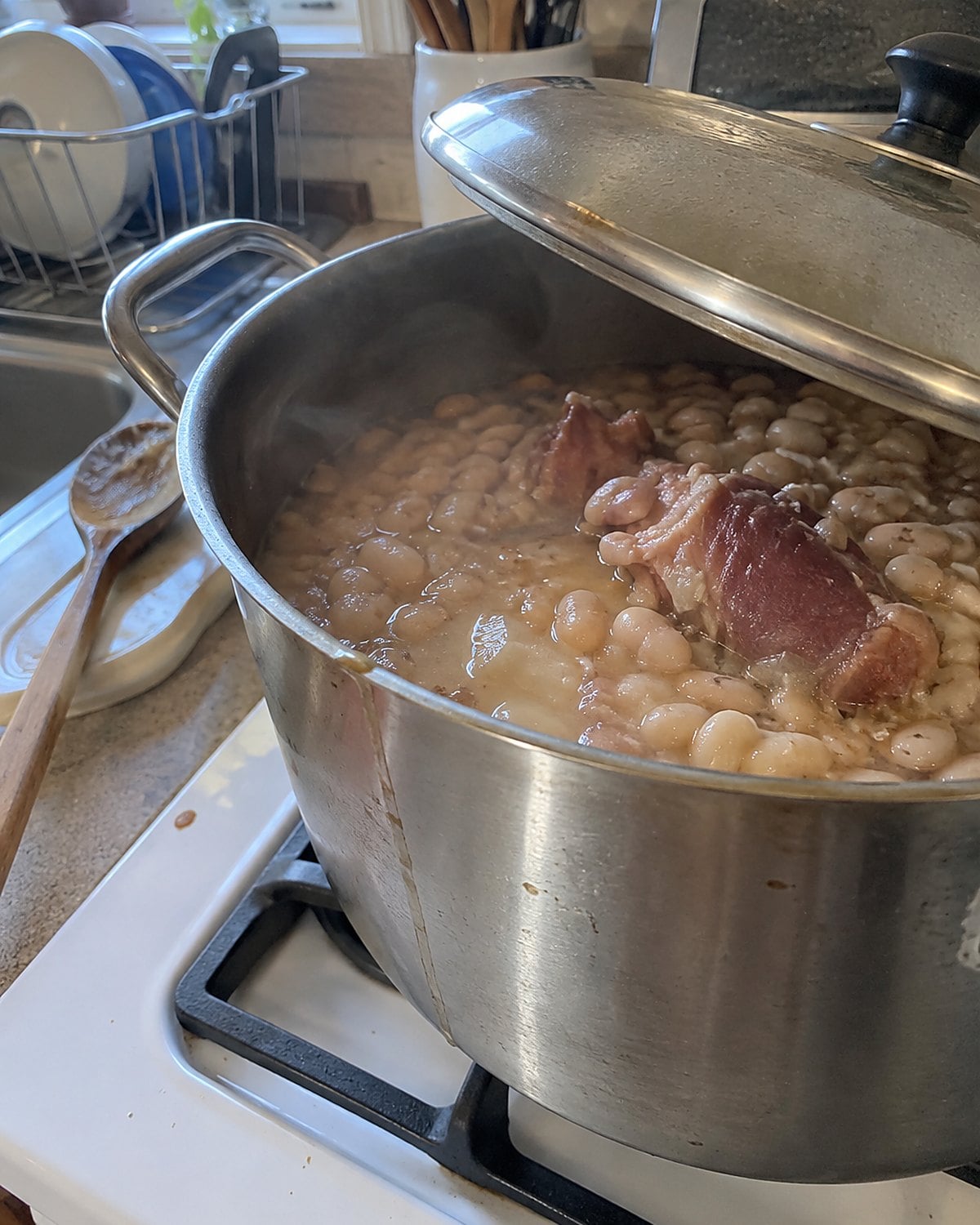 A dented pot of beans and ham hock sits on a white stove, its lid tilted open.