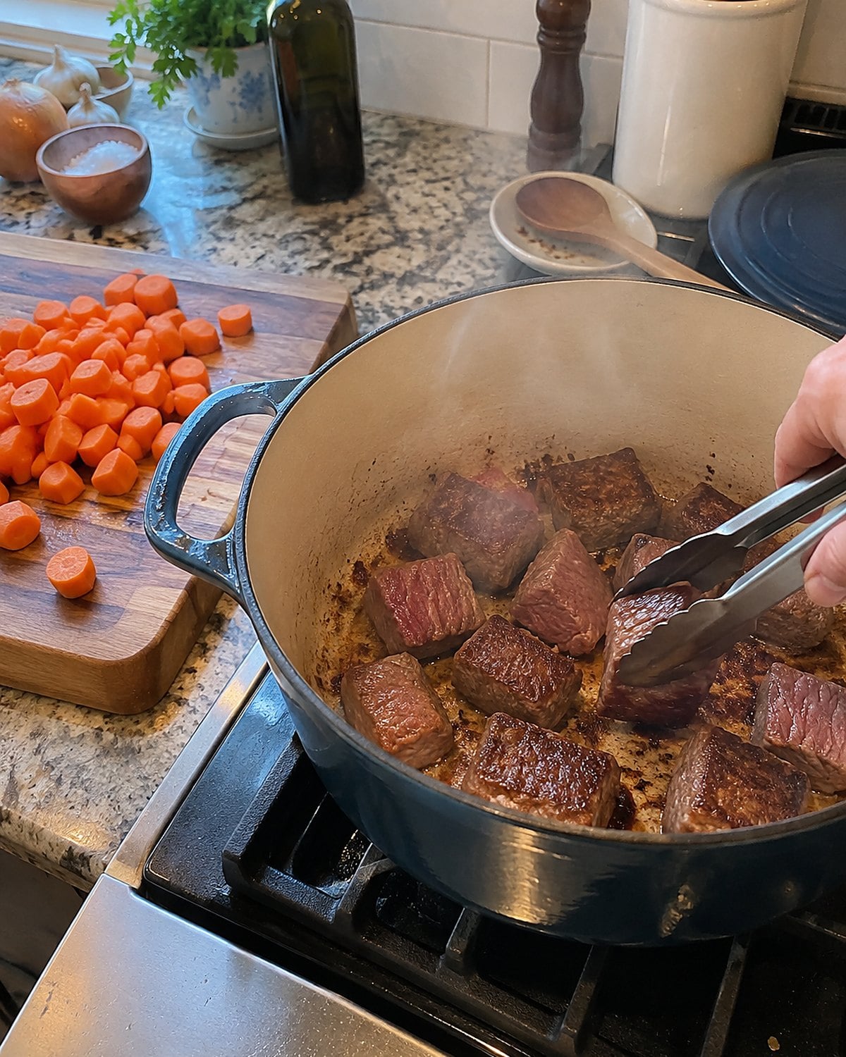 Cubes of chuck roast brown in a Dutch oven beside a board of cut carrots.