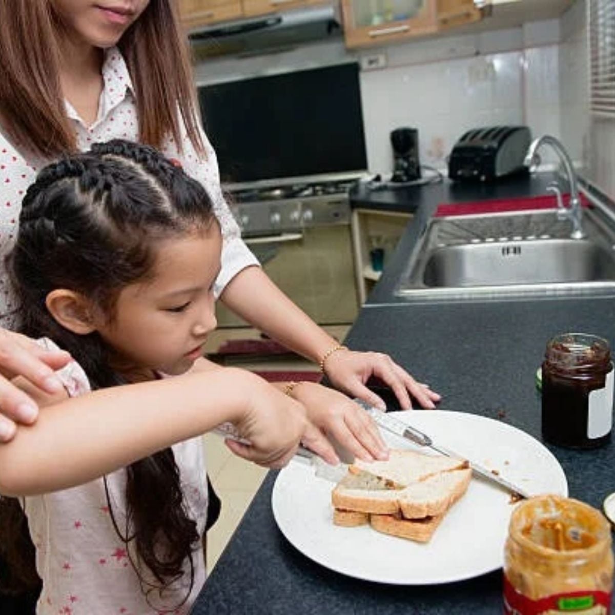 little girl cutting the peanut butter and jelly sandwich on the counter in a white plate
